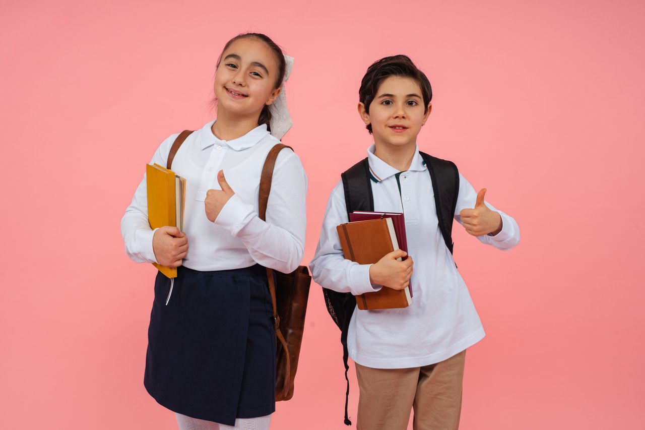 Two smiling schoolchildren give thumbs up with books and backpacks against a vibrant pink background.